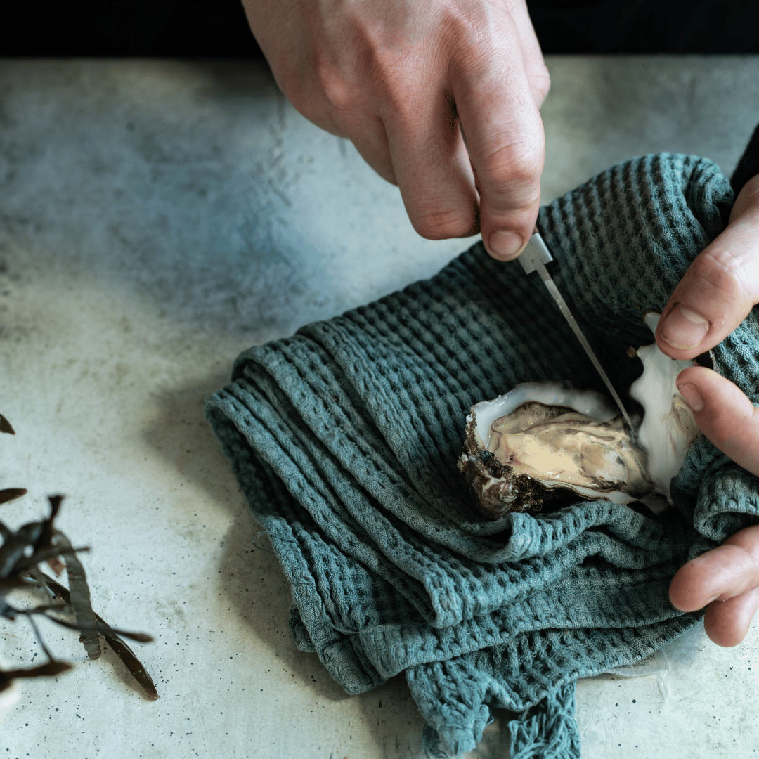 A close-up lifestyle shot of a person's hands using a knife to open an oyster, resting it on a textured teal-green waffle-knit towel over a light gray stone surface.
