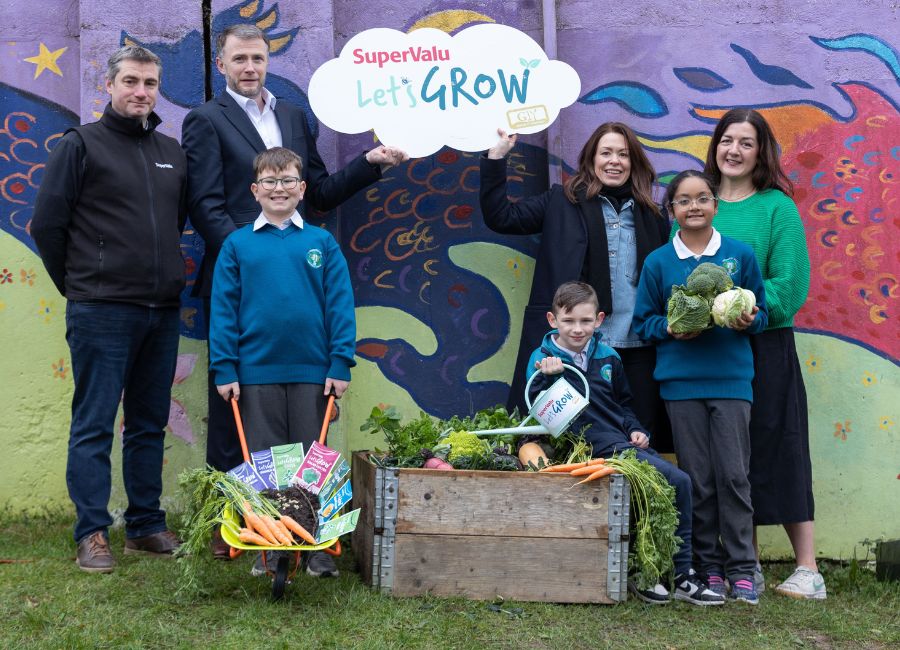 A group of 3 school children and 4 adults stand in front of a raised bed and hold up a cloud shaped sign that says SuperValu Let's GROW