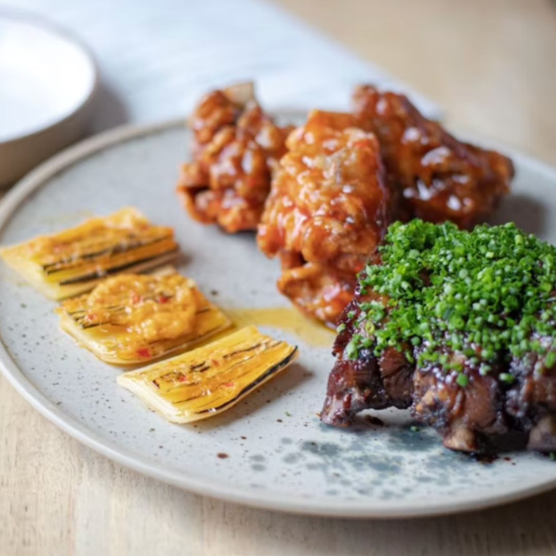 Filipino tasting plate featuring glazed chicken wings, grilled plantains, and beef short ribs topped with chopped chives, created by Bahay.