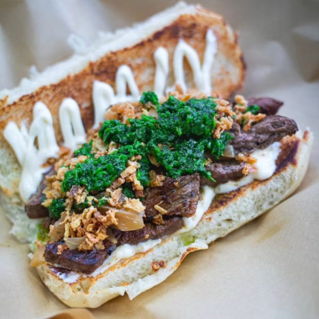 Close-up of a Filipino-style beef sandwich from Bahay, topped with crispy onions, green herb sauce, and mayonnaise on toasted bread.