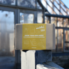 A GROWBox labeled 'GROW YOUR OWN HERBS' placed on a fence outside with a greenhouse in the background.