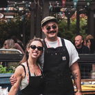 Richie Castillo and Alex O’Neill of Bahay smiling together at a food event, wearing aprons and lanyards, surrounded by people and garden planters.